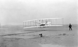 Orville fliegt in Kitty Hawk. Die Brüder Wright bauen das erste gesteuerte Motorflugzeug. By John T. Daniels [Public domain], via Wikimedia Commons